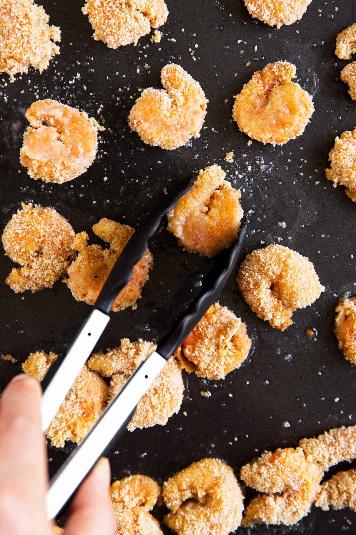 female hand using kitchen tongs to flip breaded shrimp on dark sheet pan