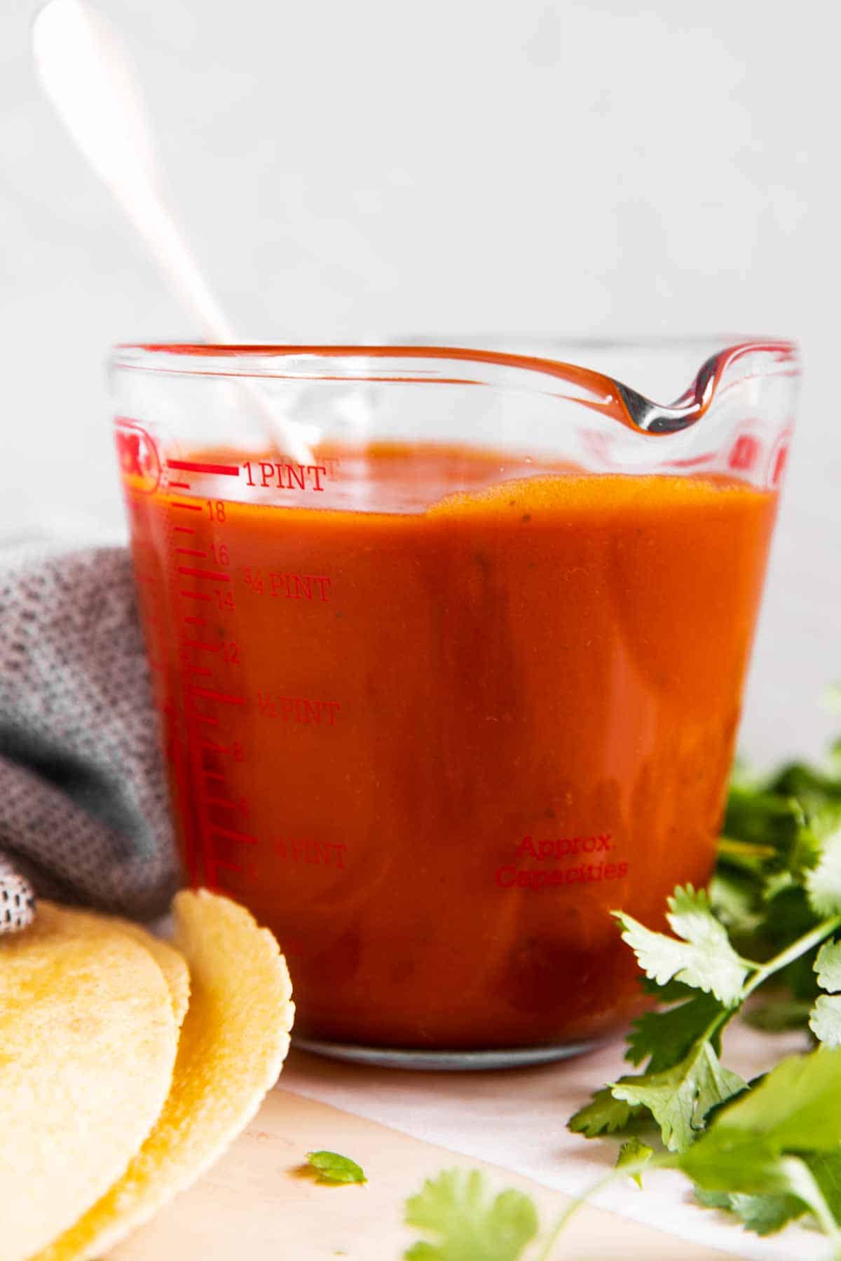 glass measuring jug with enchilada sauce surrounded by cilantro and corn tortillas