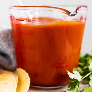 glass measuring jug with enchilada sauce surrounded by cilantro and corn tortillas