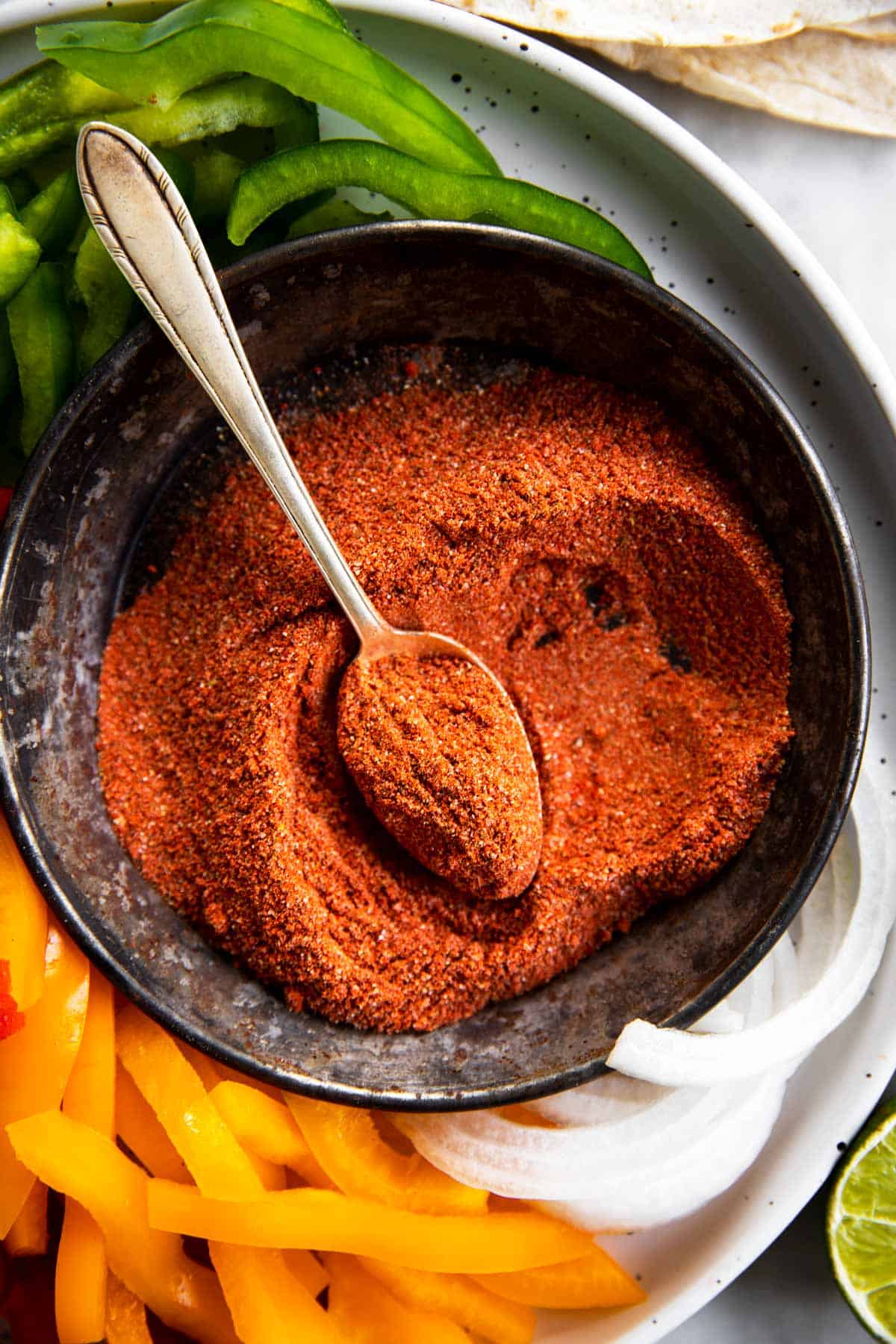 overhead view of homemade fajita seasoning in black bowl with spoon, surrounded by sliced peppers and onions