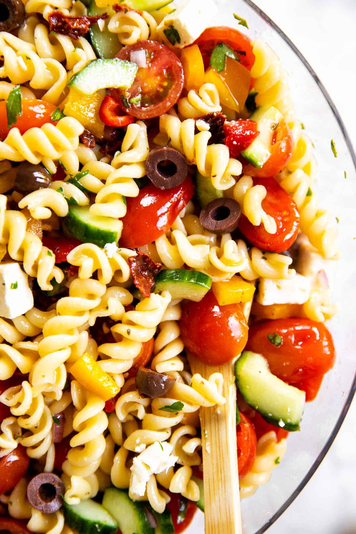 close up photo of greek pasta salad in glass bowl with wooden spoon