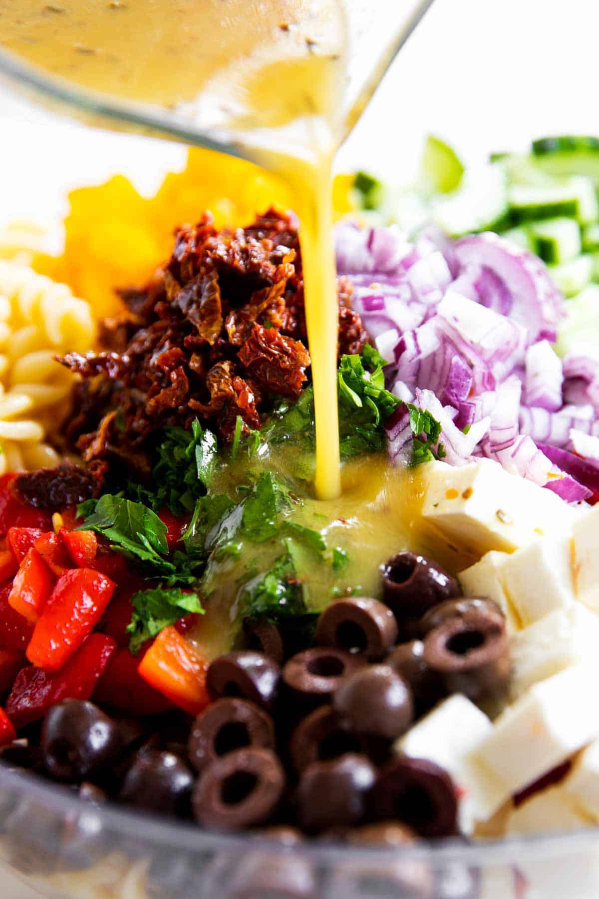 Greek salad dressing pouring over salad ingredients for Greek pasta salad