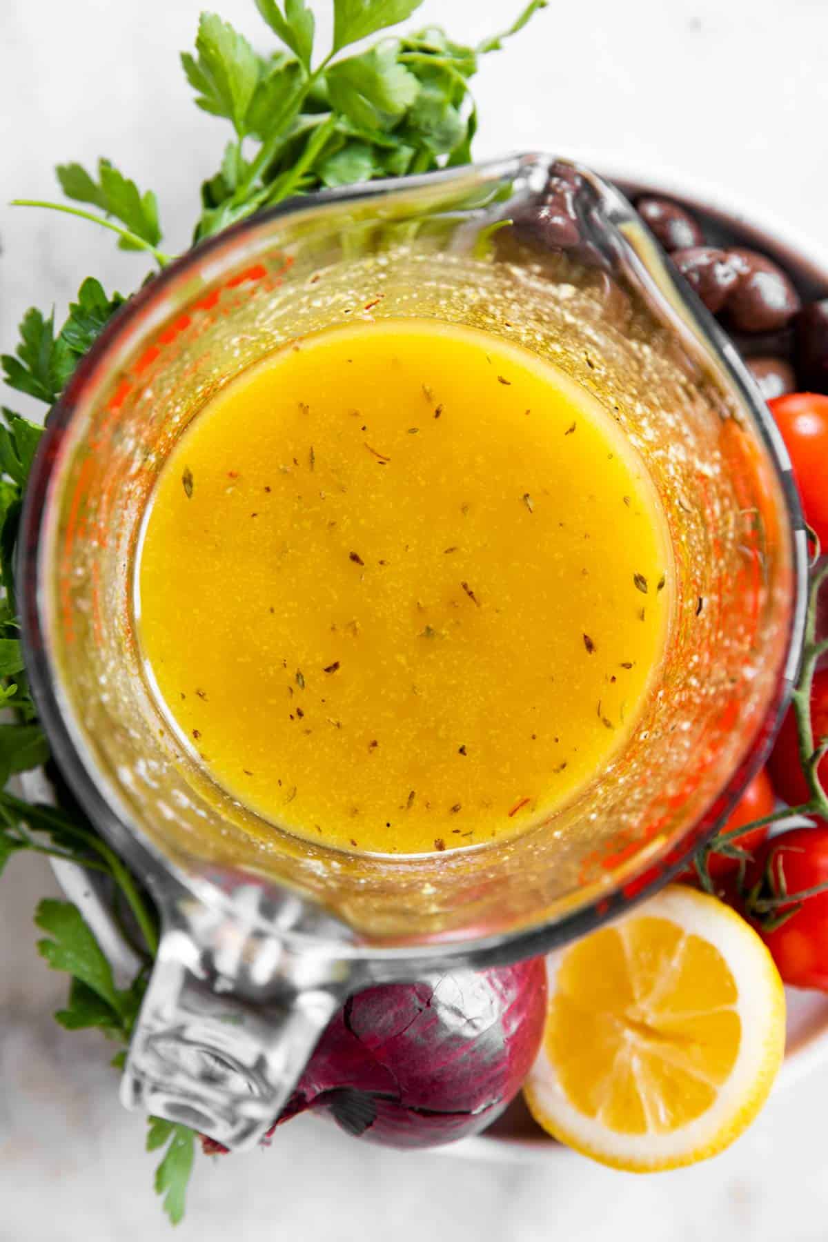 overhead view of greek salad dressing in glass measuring jug amidst fresh parsley, olives, tomatoes, red onion and halved lemon