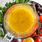 overhead view of greek salad dressing in glass measuring jug amidst fresh parsley, olives, tomatoes, red onion and halved lemon