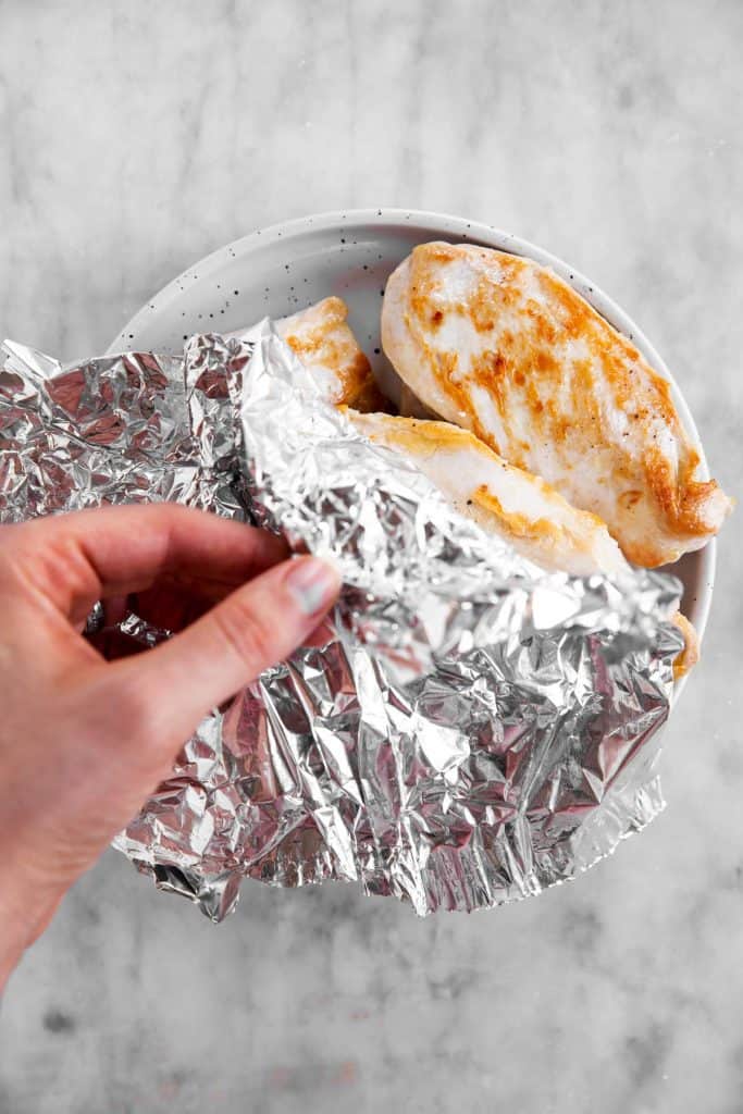 female hand covering seared chicken breast with foil on white plate