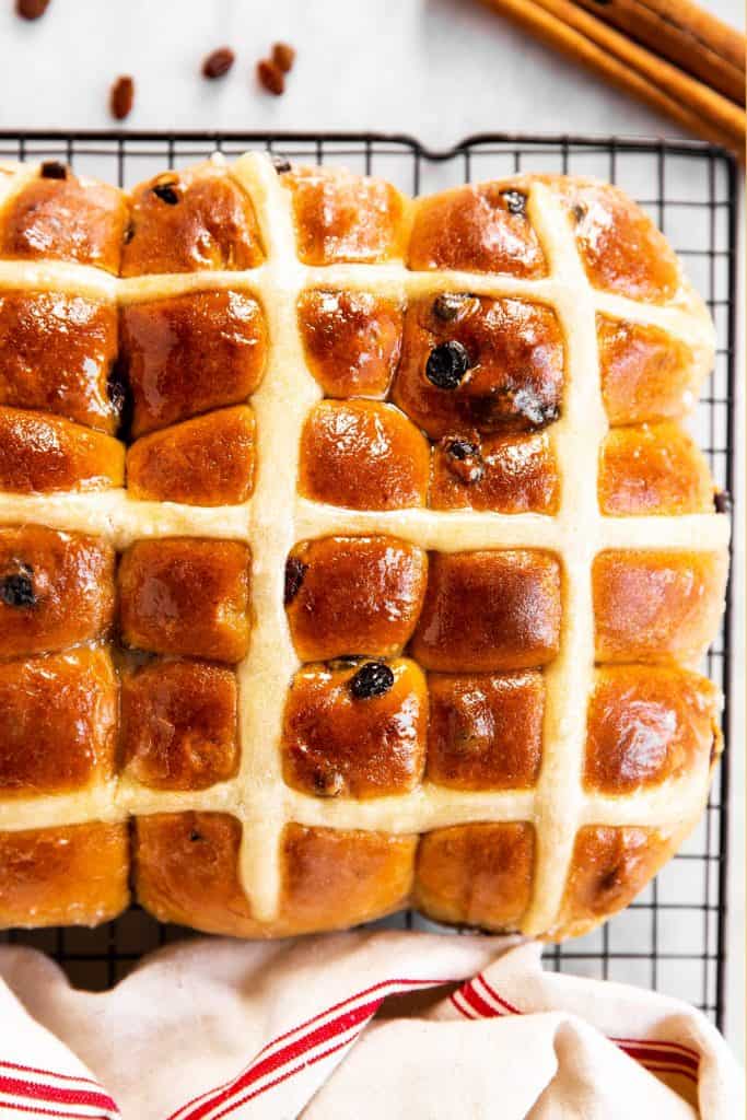 overhead view of hot cross buns on wire rack surrounded by a tea towel, raisins and cinnamons sticks