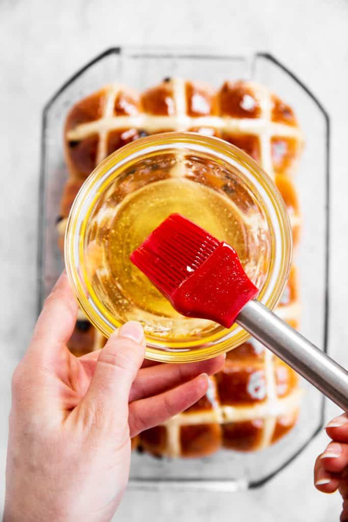 red pastry brush dipping into orange glaze over pan with hot cross buns