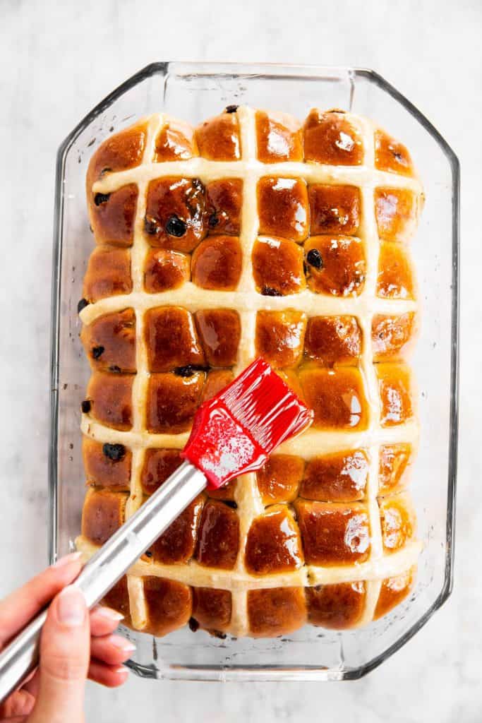 red pastry brush brushing orange glaze over baked hot cross buns in glass pan