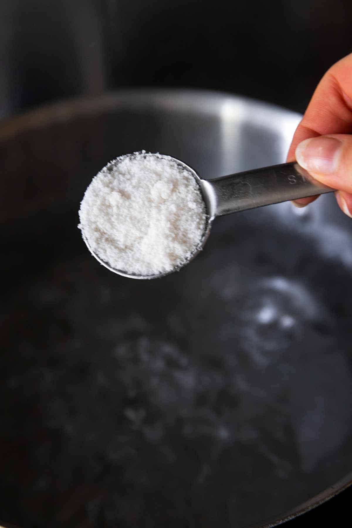 female hand holding tablespoon filled with salt over pot of boiling water