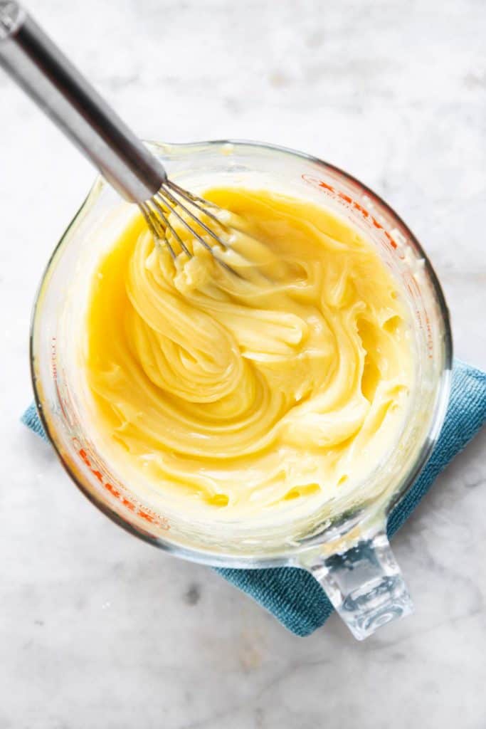 homemade mayonnaise in glass measuring jug with whisk sitting on blue cloth