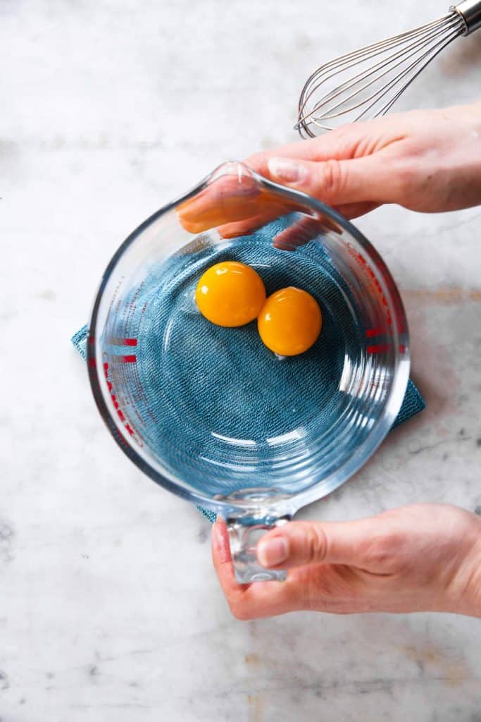 female hands placing glass measuring jug containing two egg yolks on blue cloth on marble surface