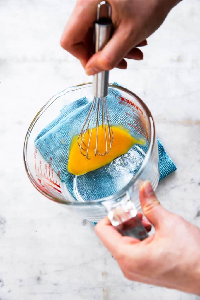 female hand whisking egg yolks in glass measuring jug sitting on top of a blue cloth