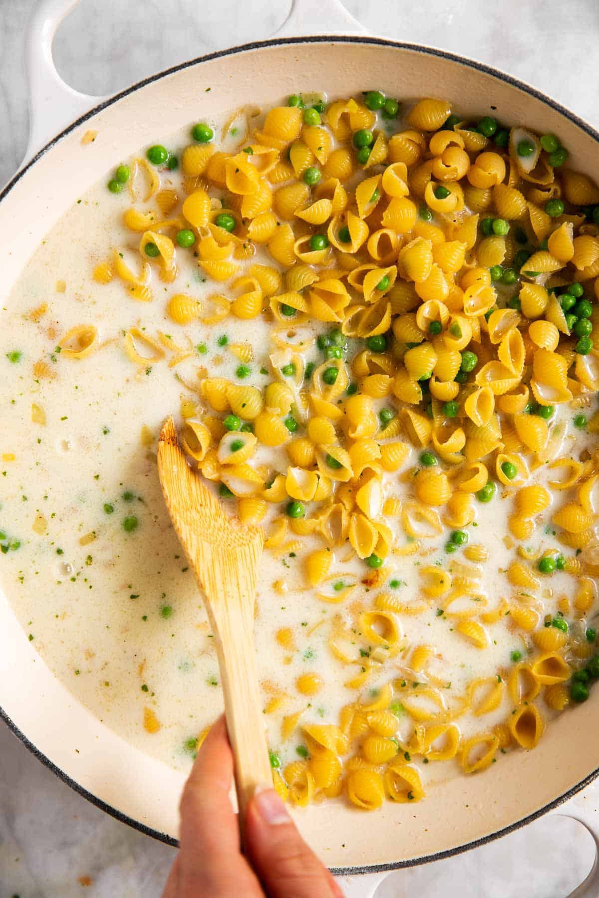 female hand stirring pasta and peas into milk with wooden spoon