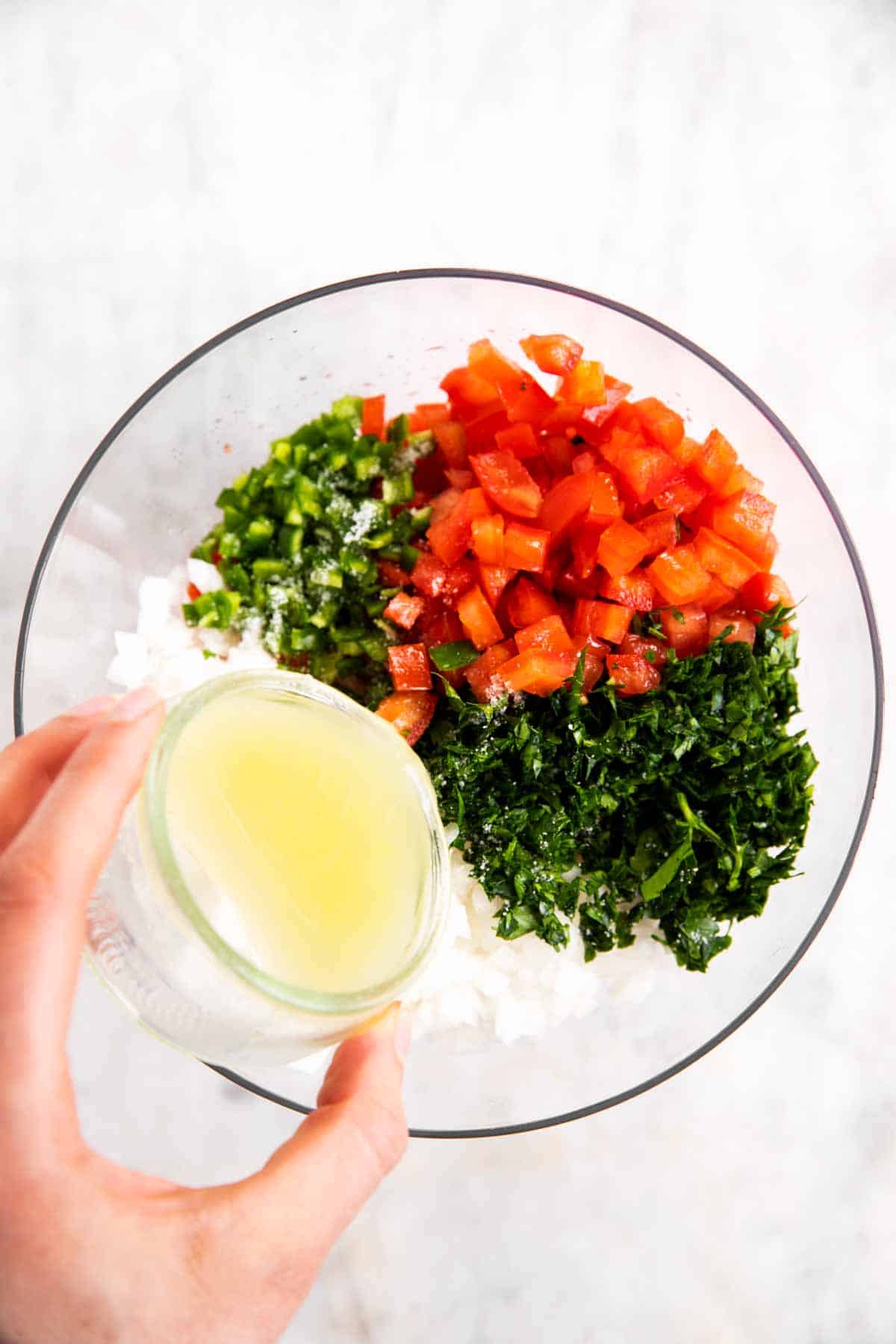 female hand pouring lime juice over ingredients for pico de Gallo in glass bowl