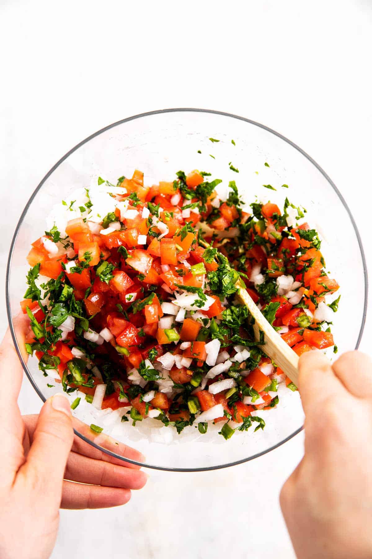 hands stirring pico de Gallo in glass bowl with wooden spoon