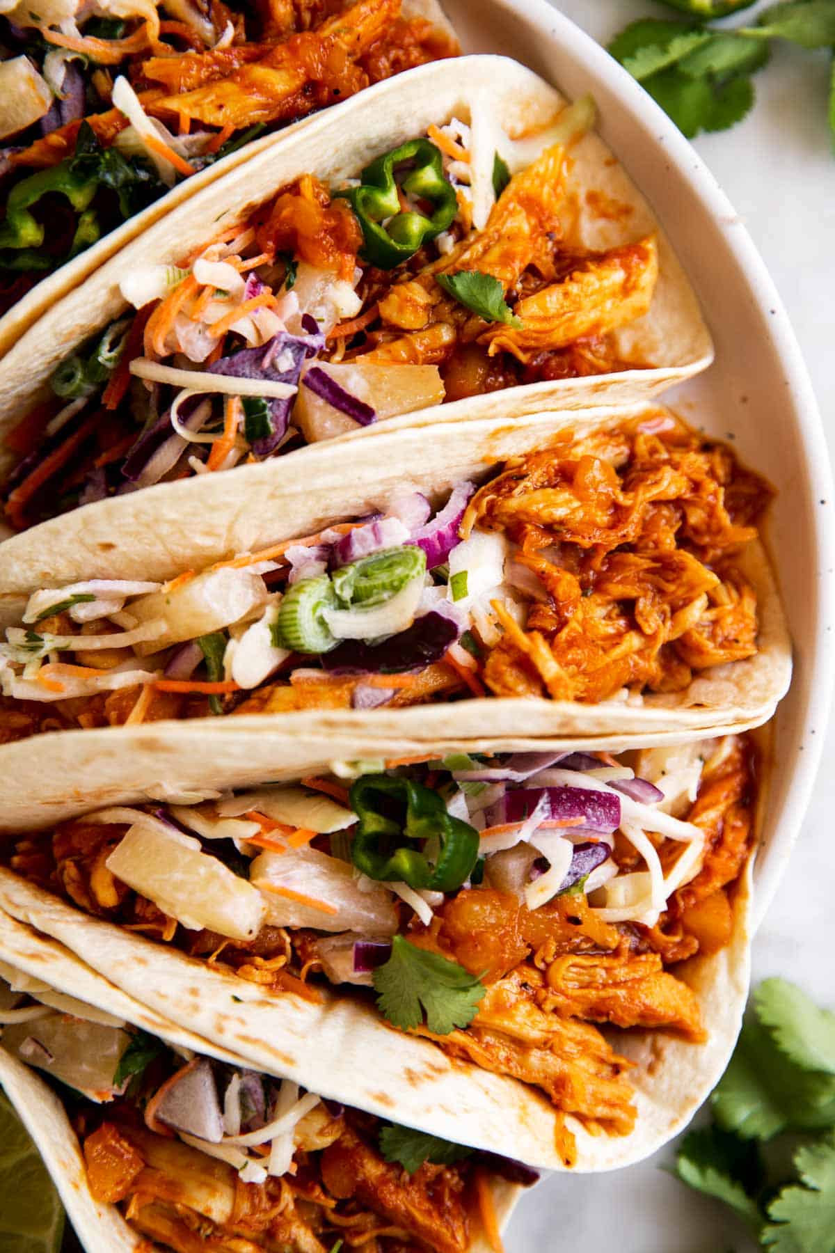 overhead view of 5 pineapple bbq pulled chicken tacos on white plate with fresh cilantro