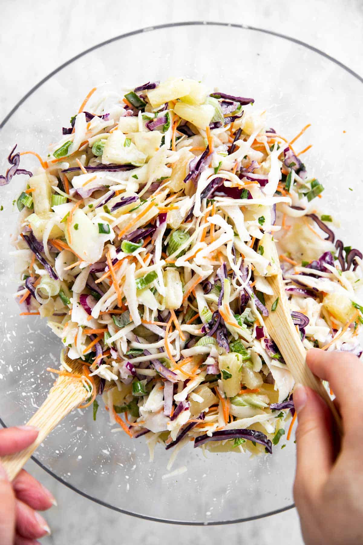 female hands using wooden salad tongs to toss pineapple coleslaw in glass bowl