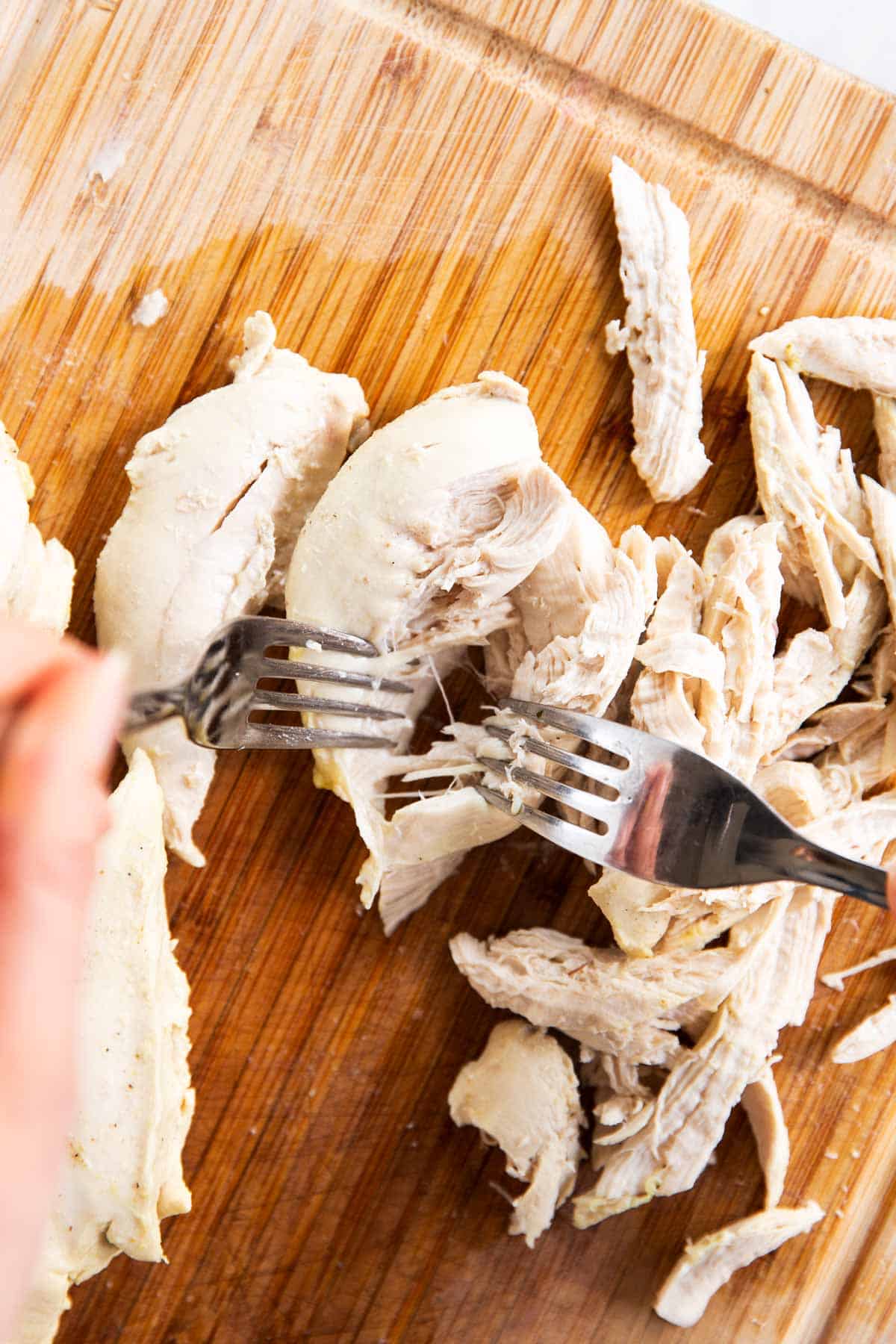 female hands using two forks to shred poached chicken breast on wooden board