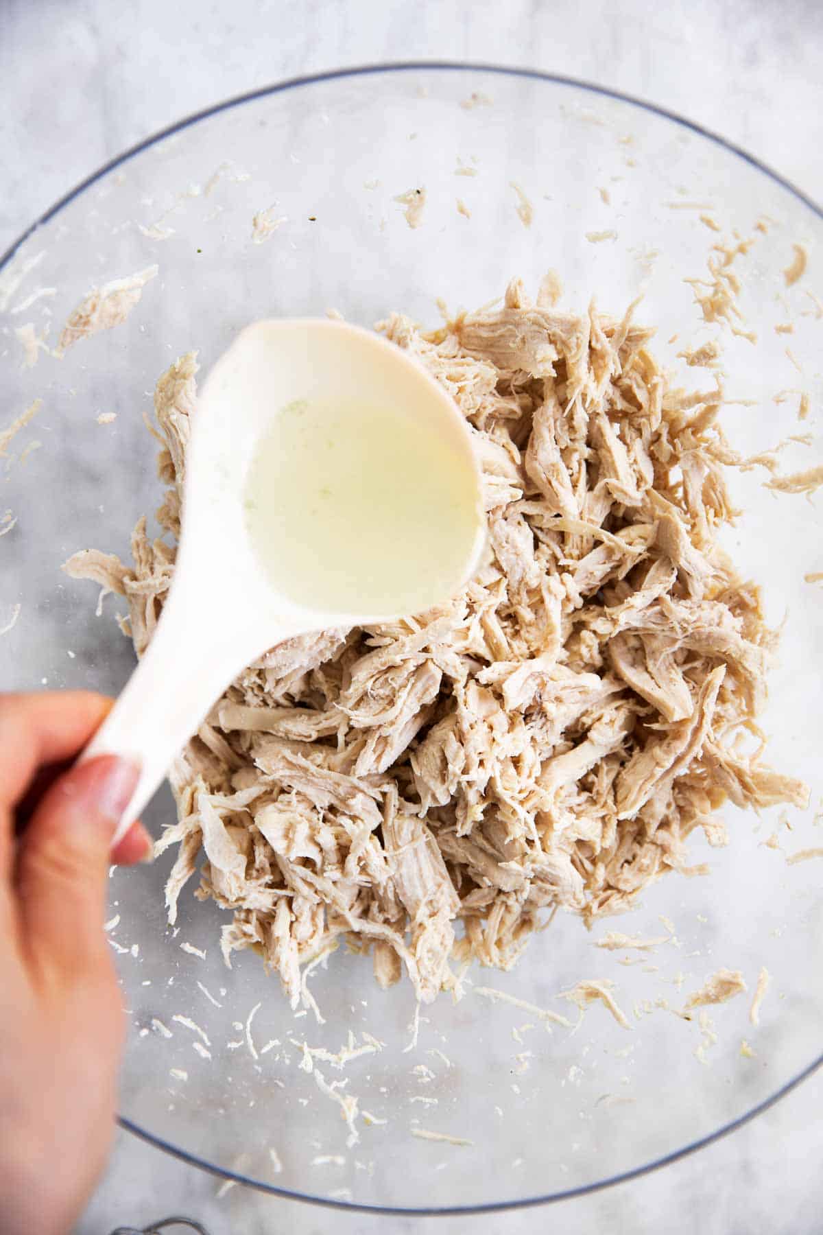 female hand using white spoon to pour broth over shredded chicken in glass bowl