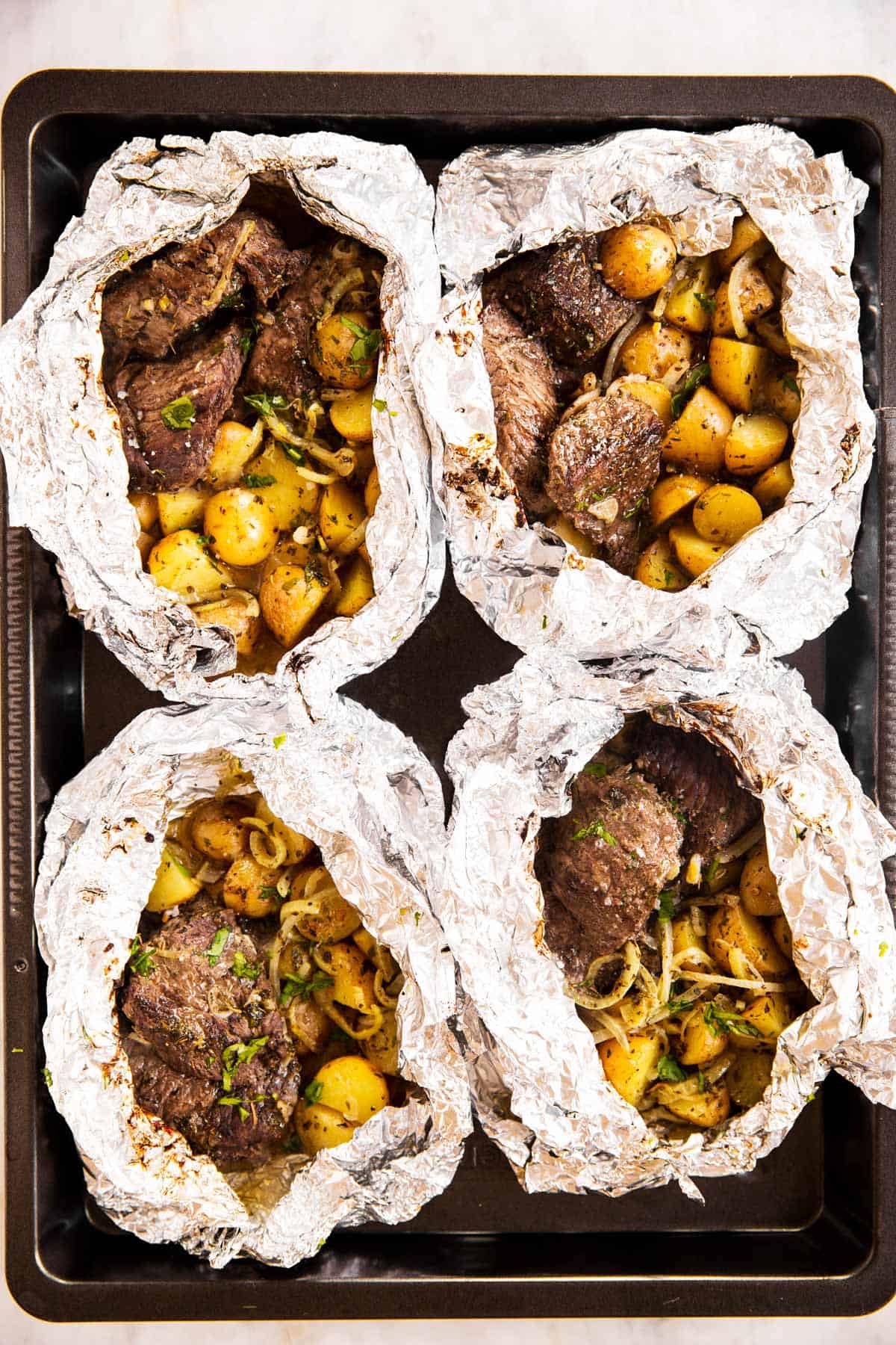 overhead view of four opened steak and potato foil packets on black sheet pan