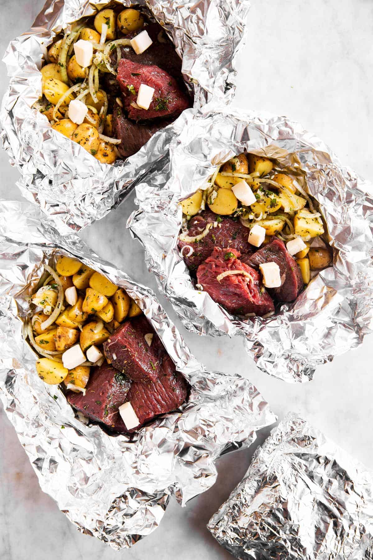 three unclosed steak and potato foil packets on marble surface