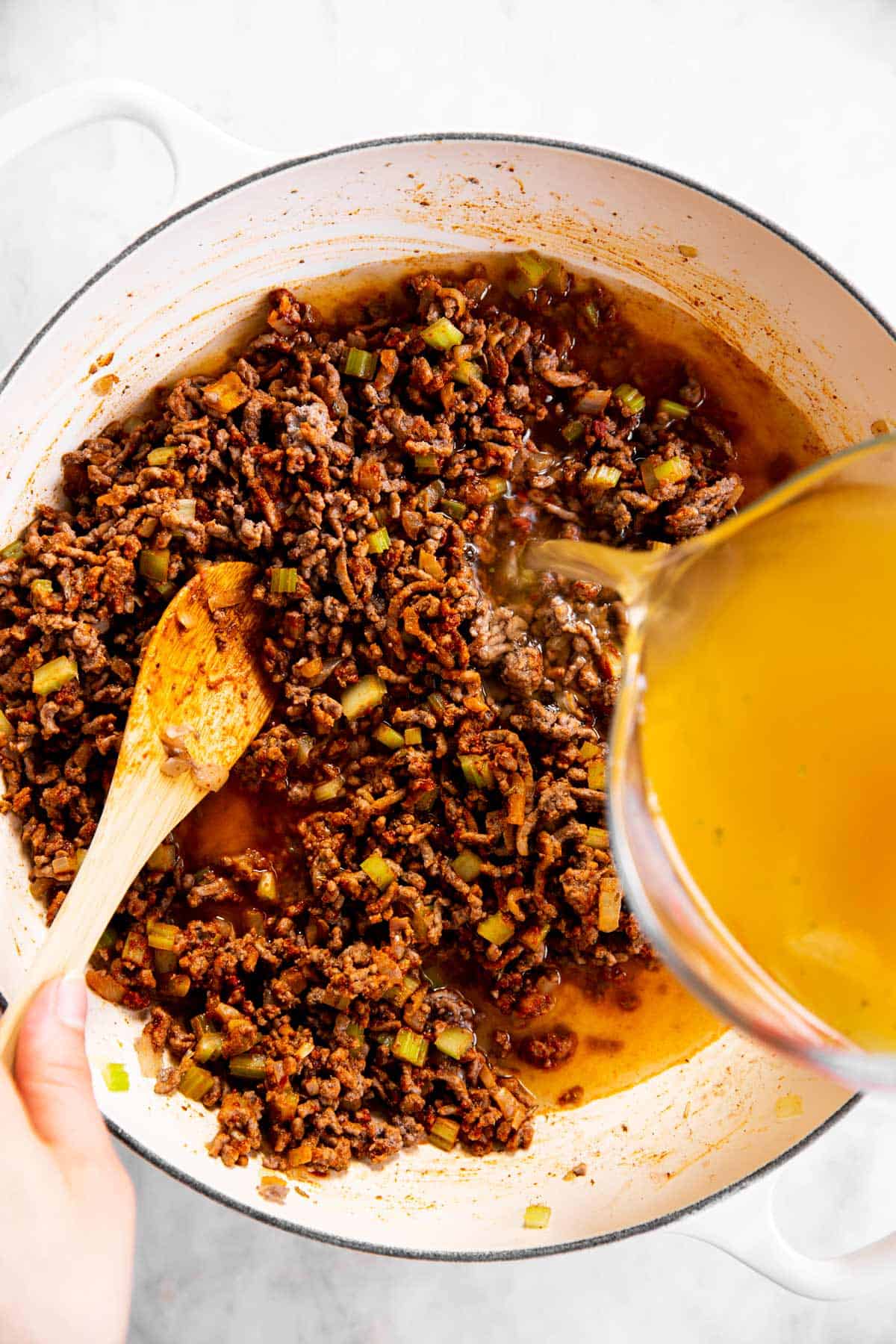 female hand pouring chicken broth into pan with browned ground beef and vegetables