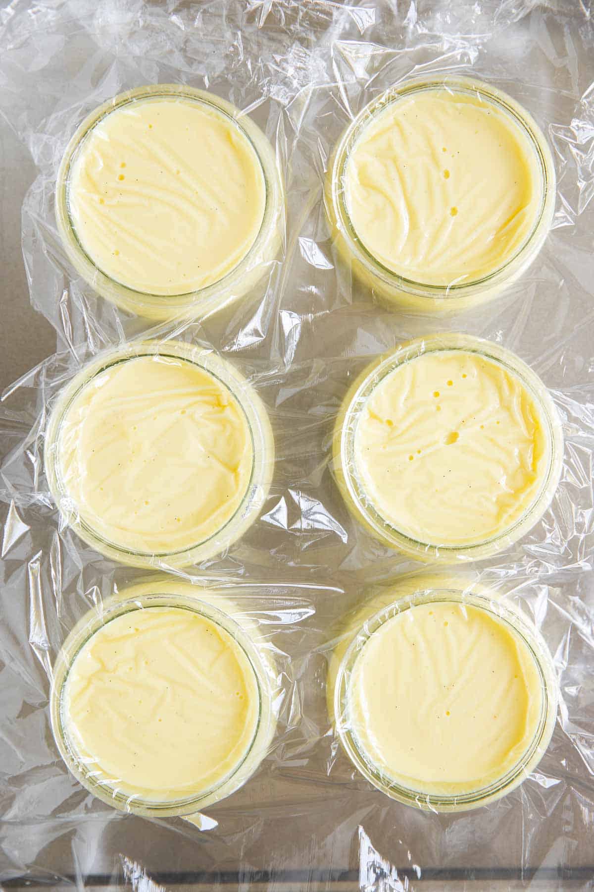 six glass jars filled with vanilla pudding and covered with plastic wrap