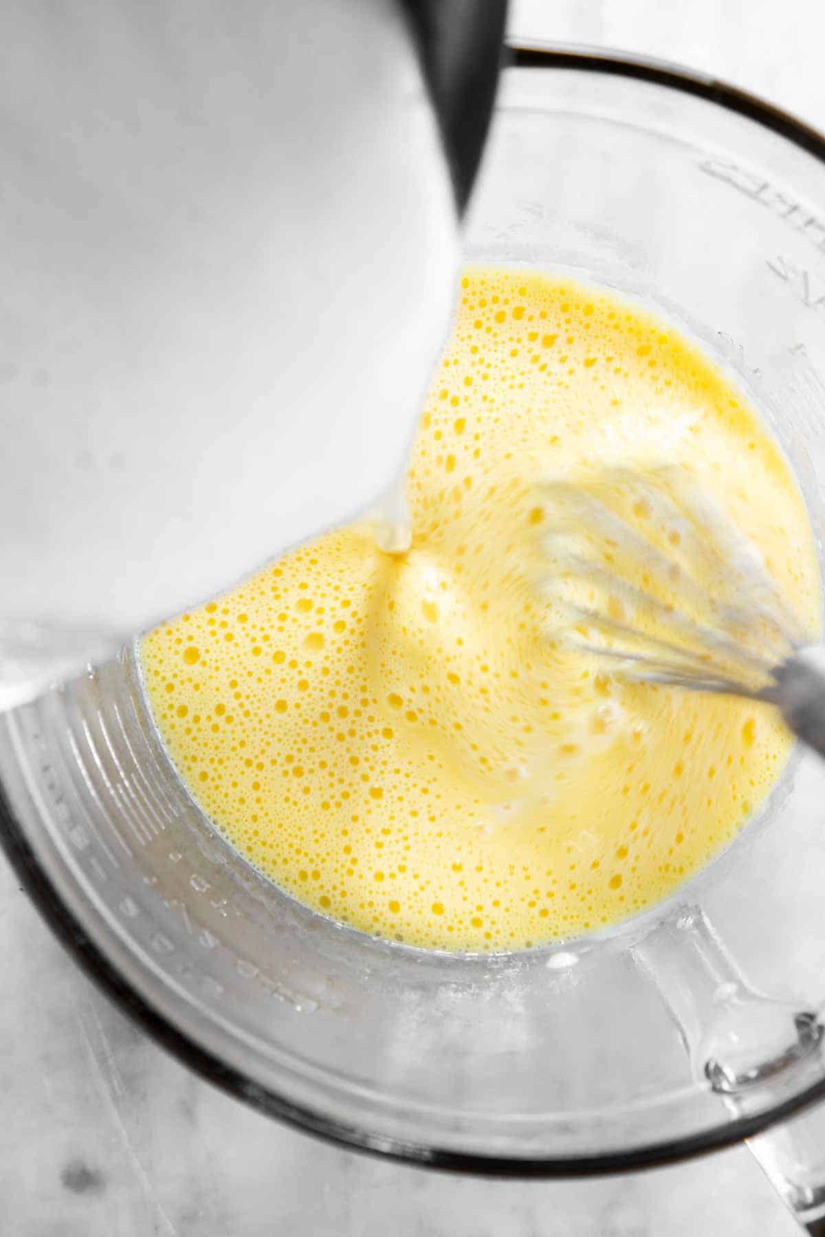 milk pouring from saucepan into glass bowl with beaten egg yolks