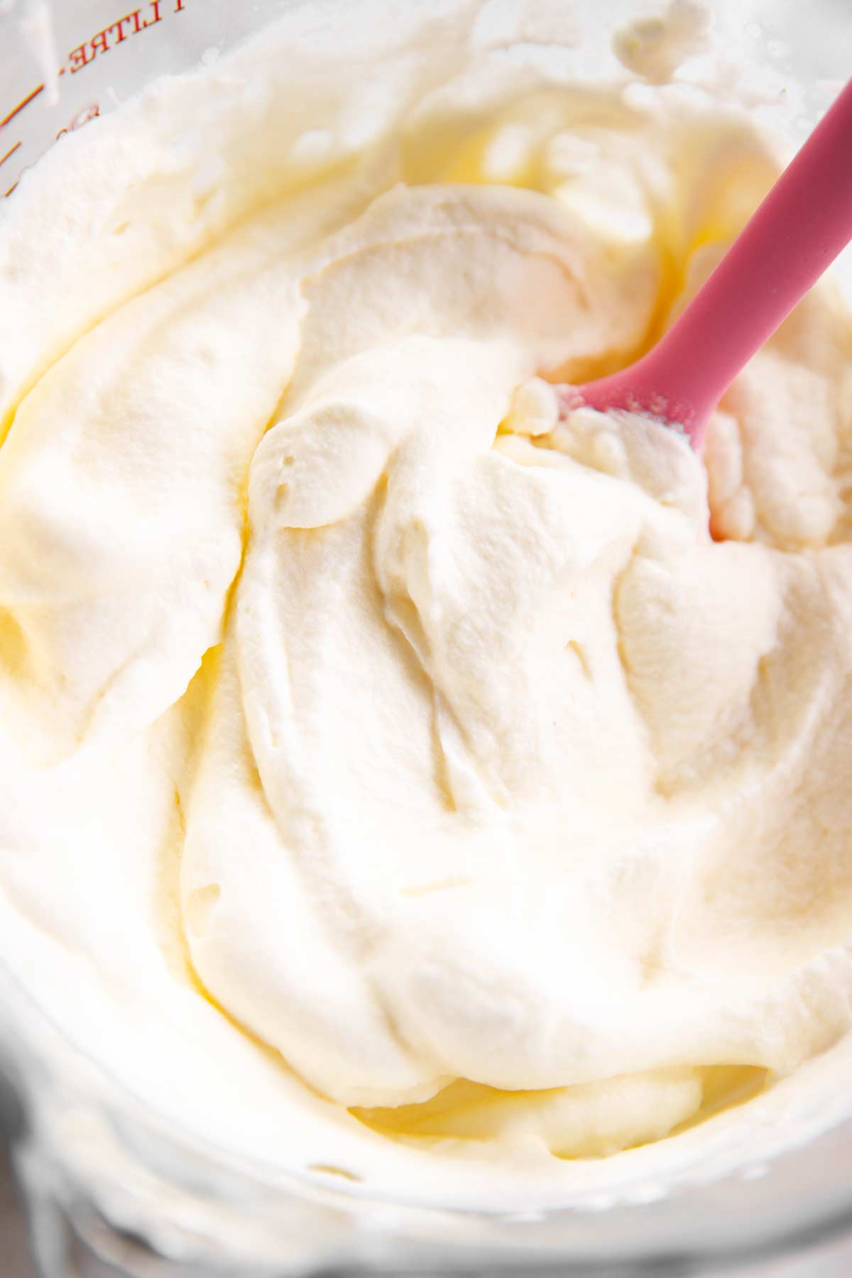 close up photo of homemade whipped cream in glass measuring jug with pink rubber spatula