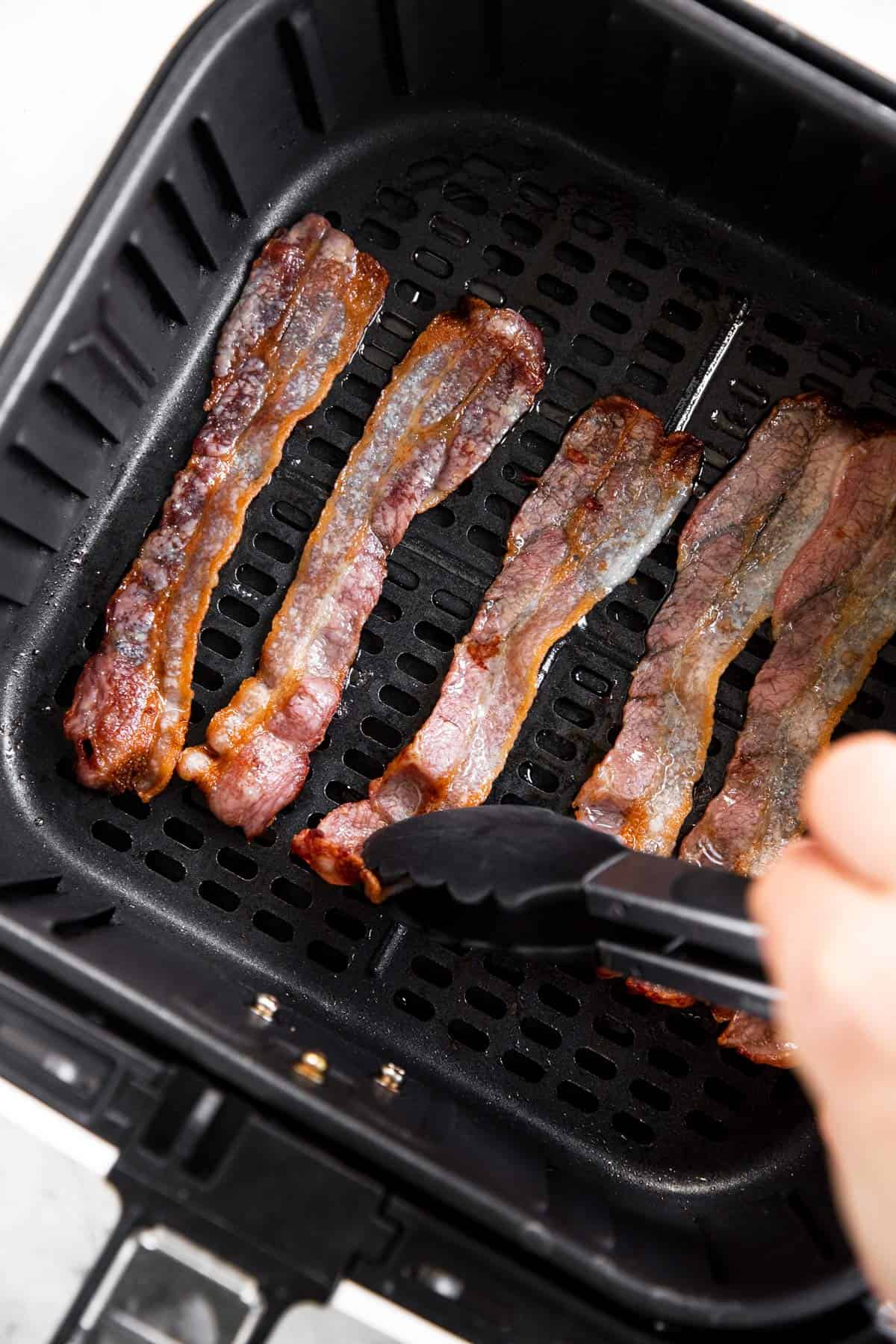 female hand using kitchen tongs to flip bacon in air fryer basket