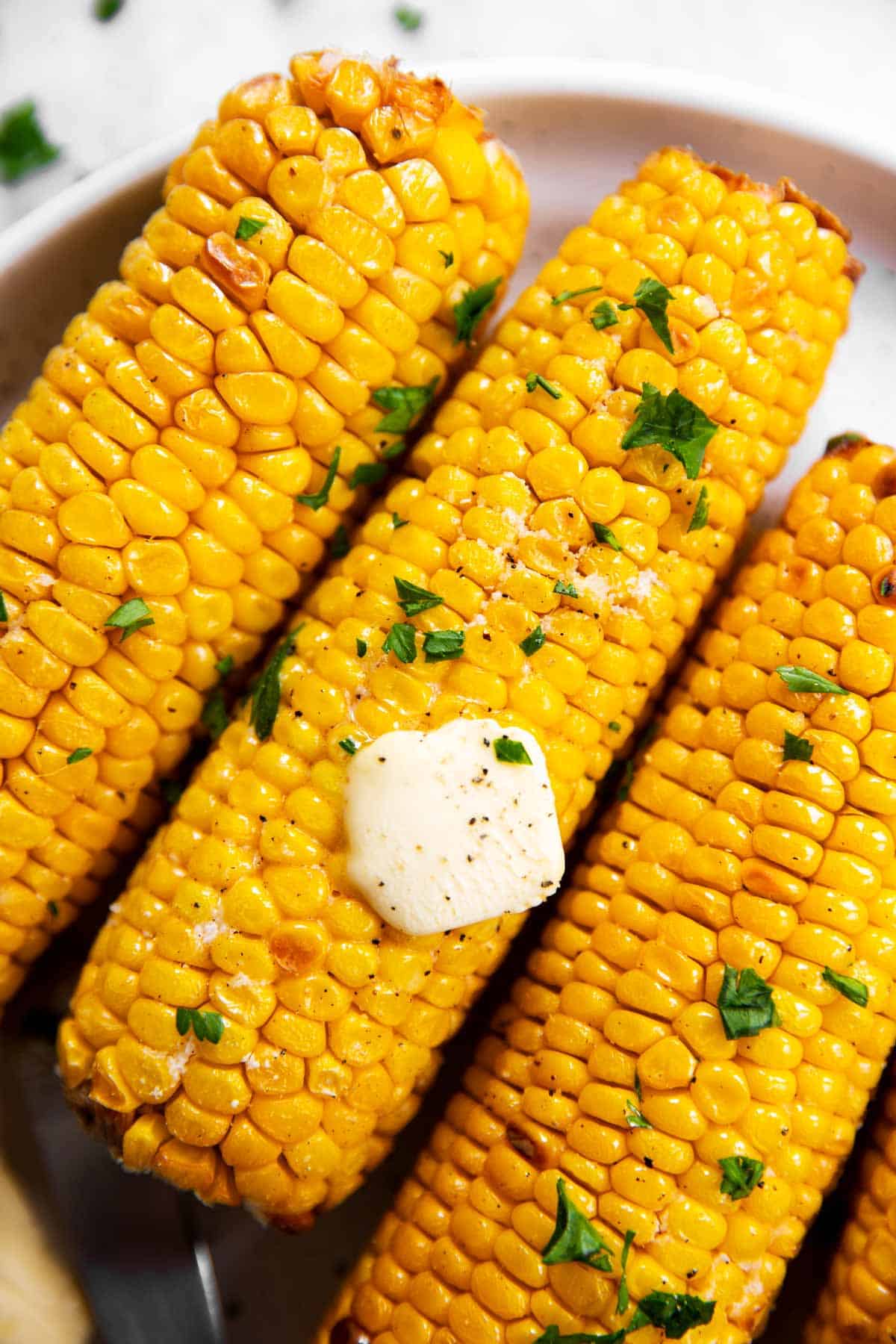 close up photo of air fried corn on the cob on a white plate, with butter and chopped parsley