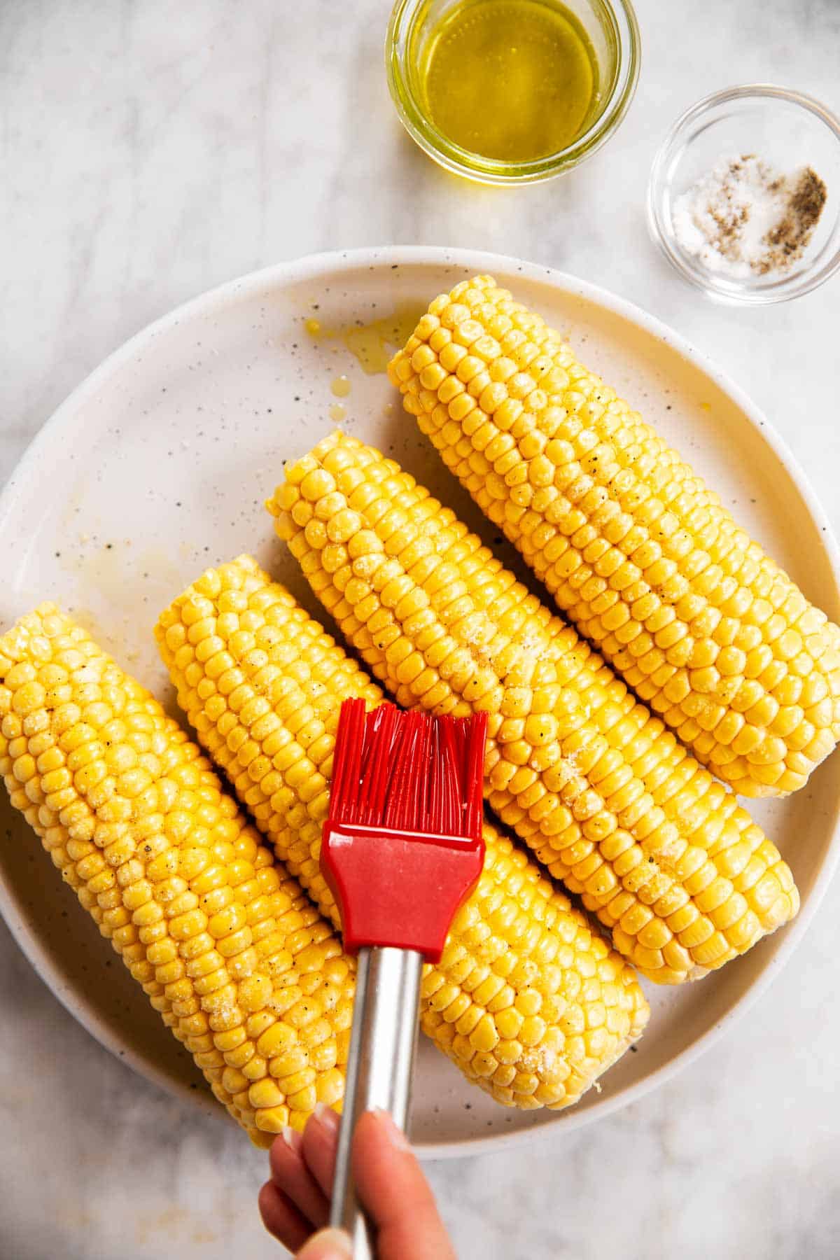 four ears of shucked corn on white plate, female hand brushing one with oil using a red pastry brush