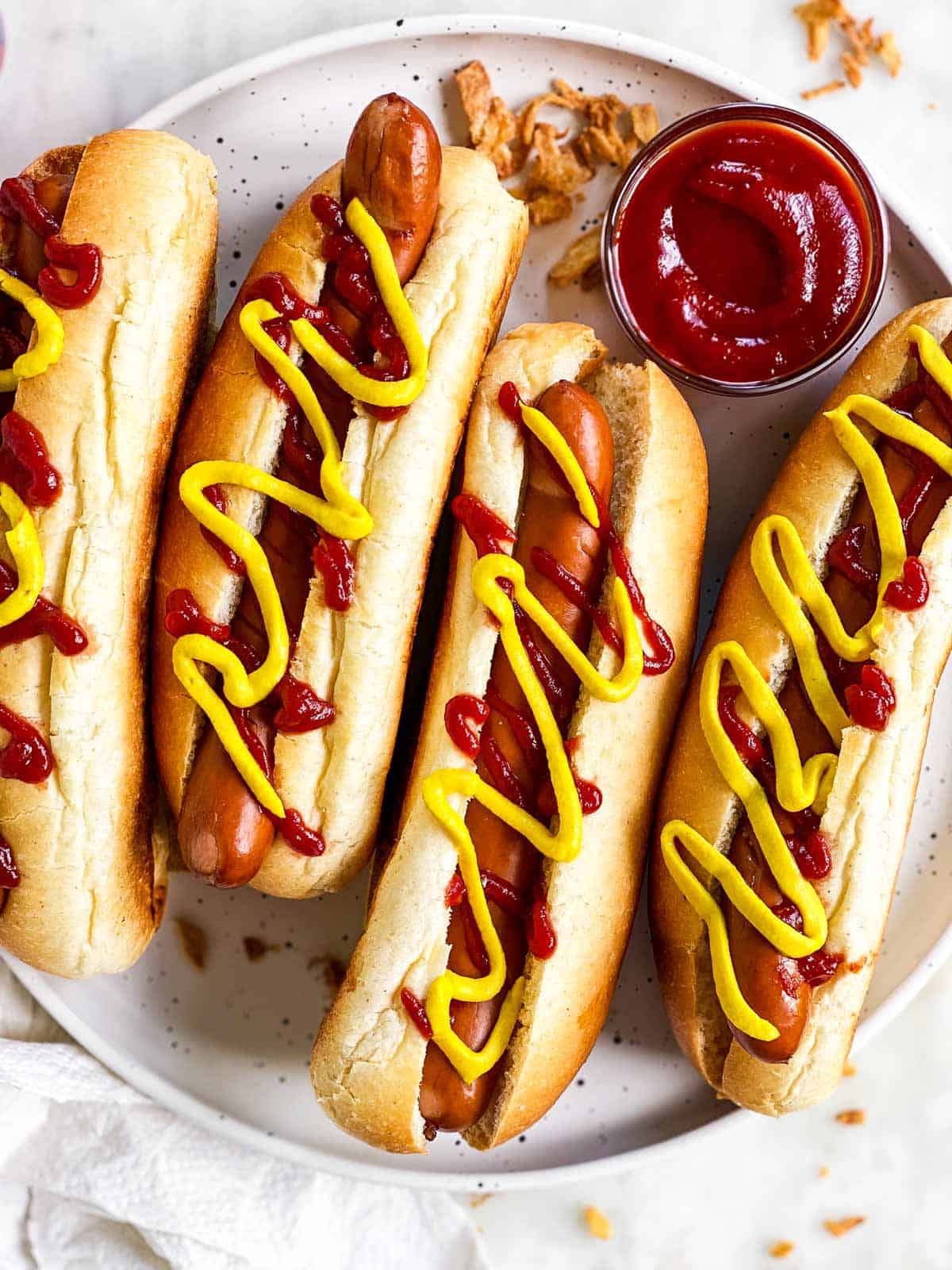 four hot dogs garnished with ketchup and mayo on white plate with more ketchup and fried onions