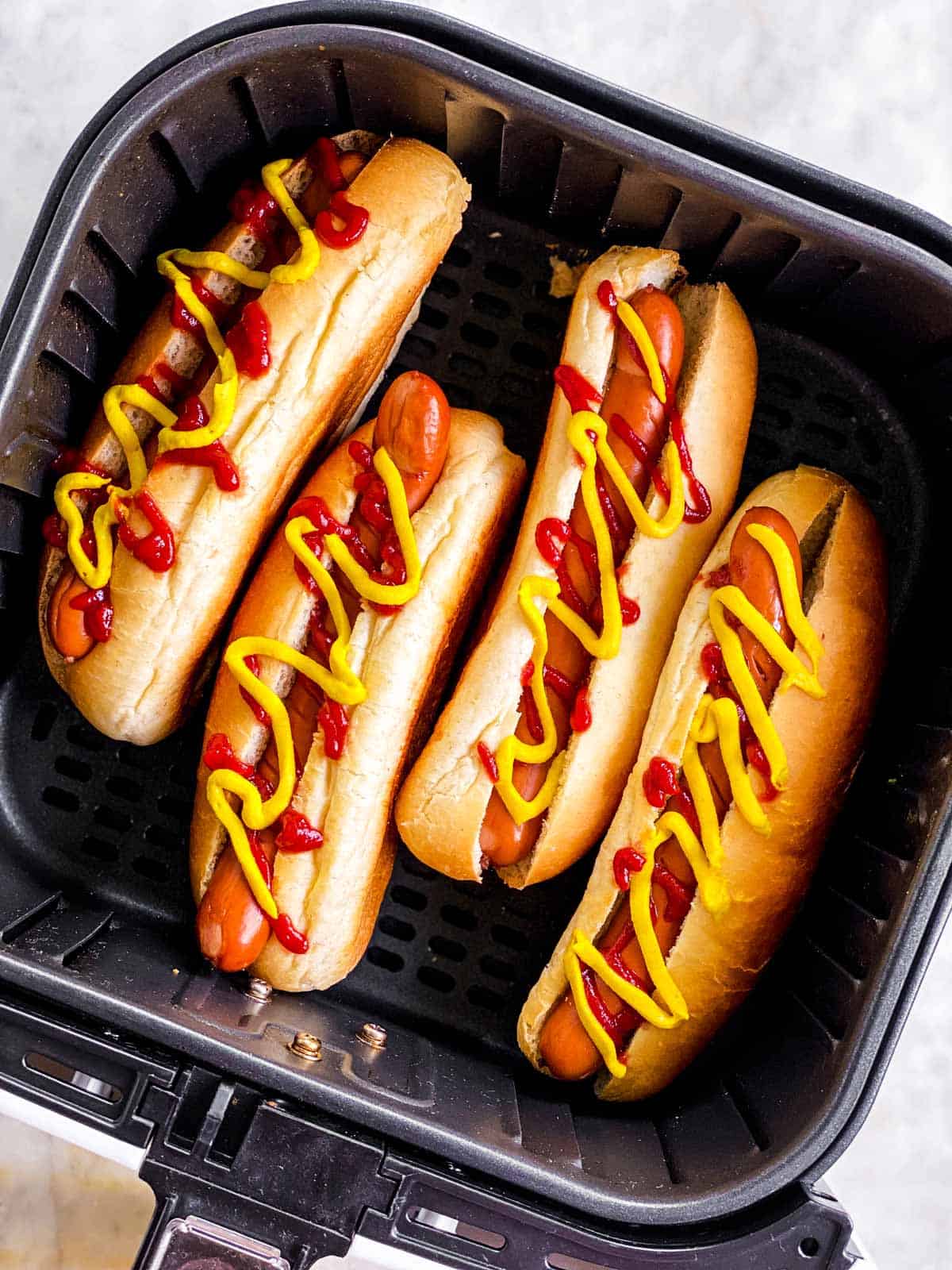 overhead view of four hot dogs garnished with ketchup and mustard in square air fryer basket