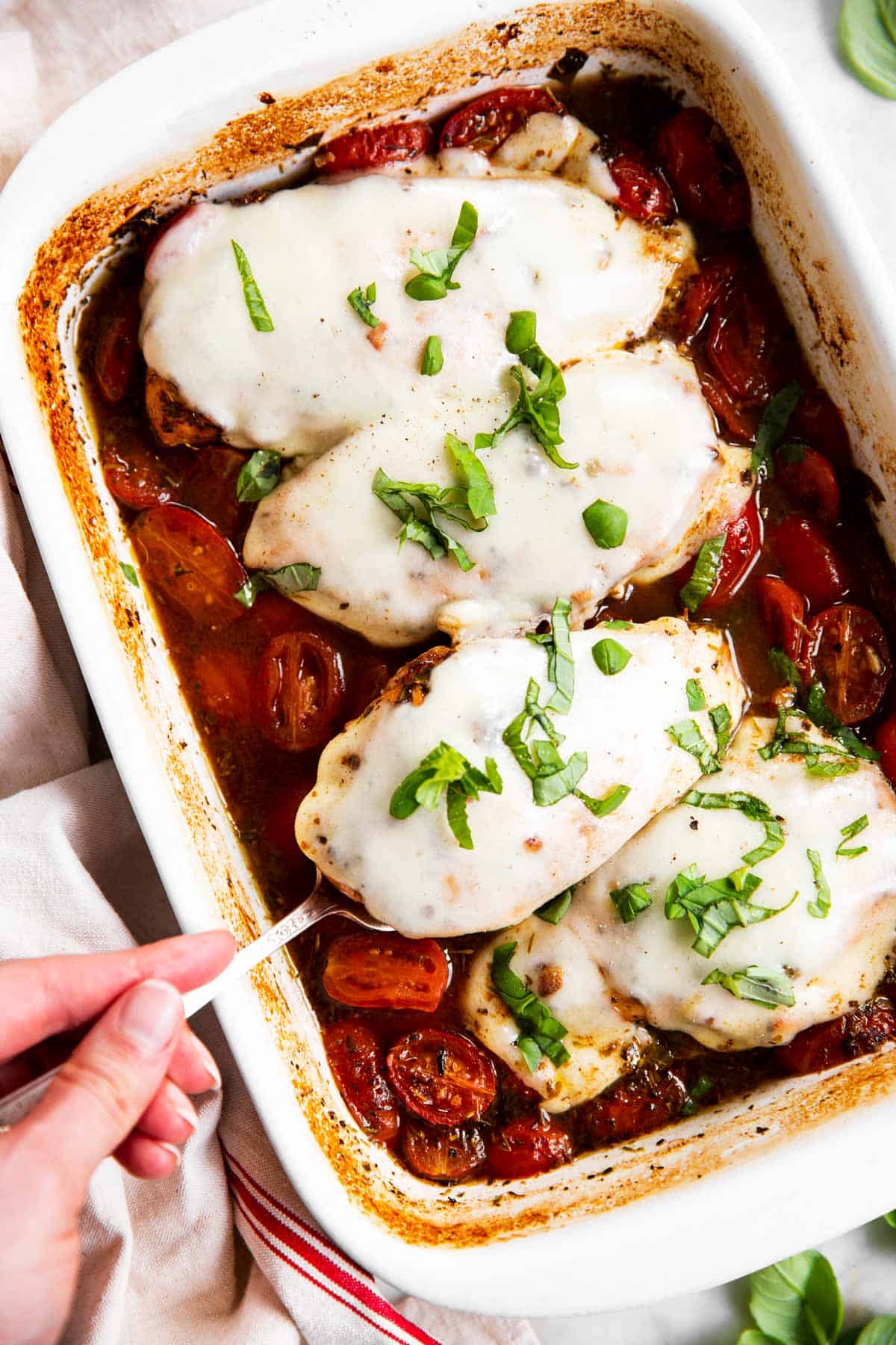 female hand using serving fork to remove a chicken breast from a dish of baked caprese chicken 