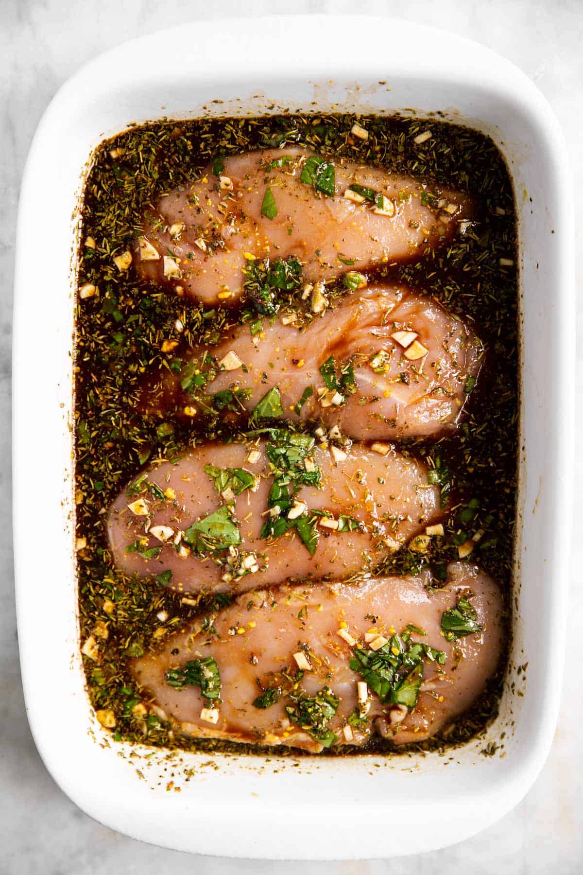 four raw chicken breasts in white casserole dish with balsamic marinade