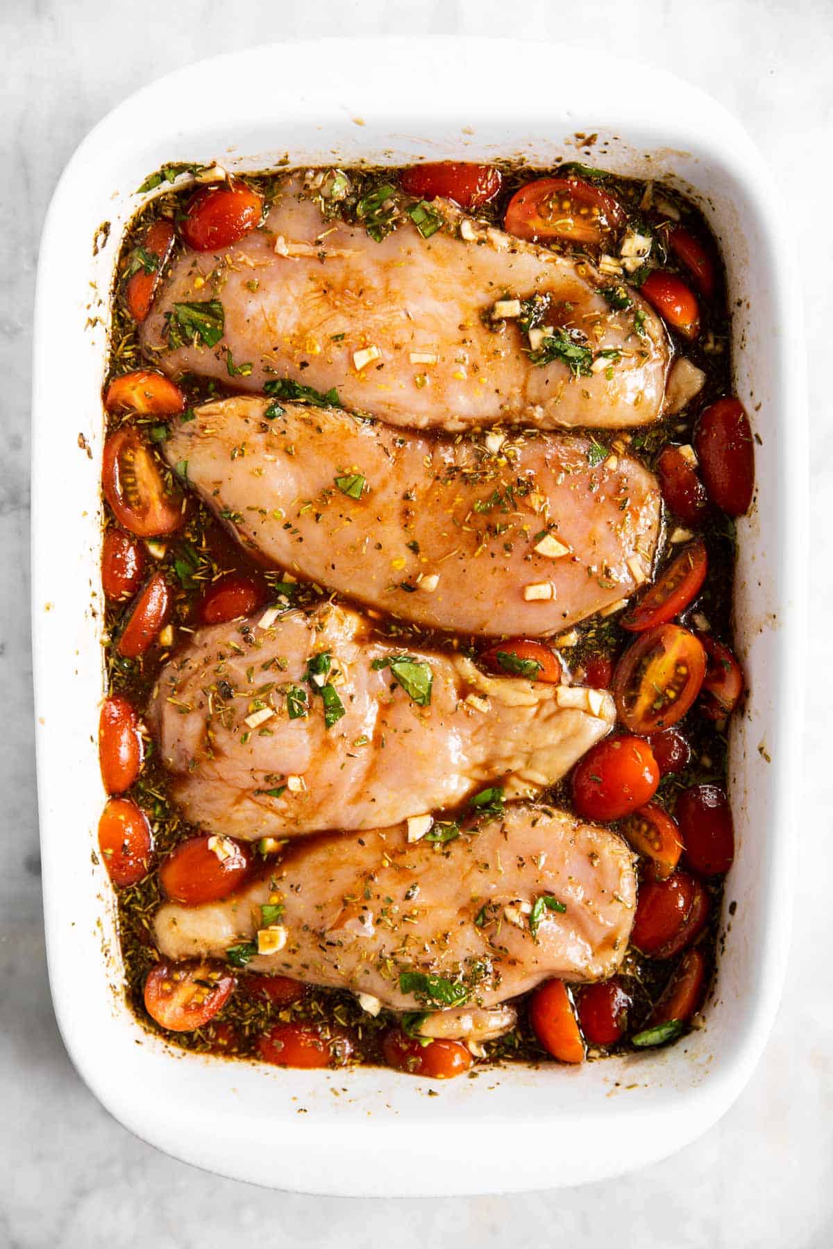 four raw chicken breasts in white casserole dish with balsamic marinade and halved cherry tomatoes