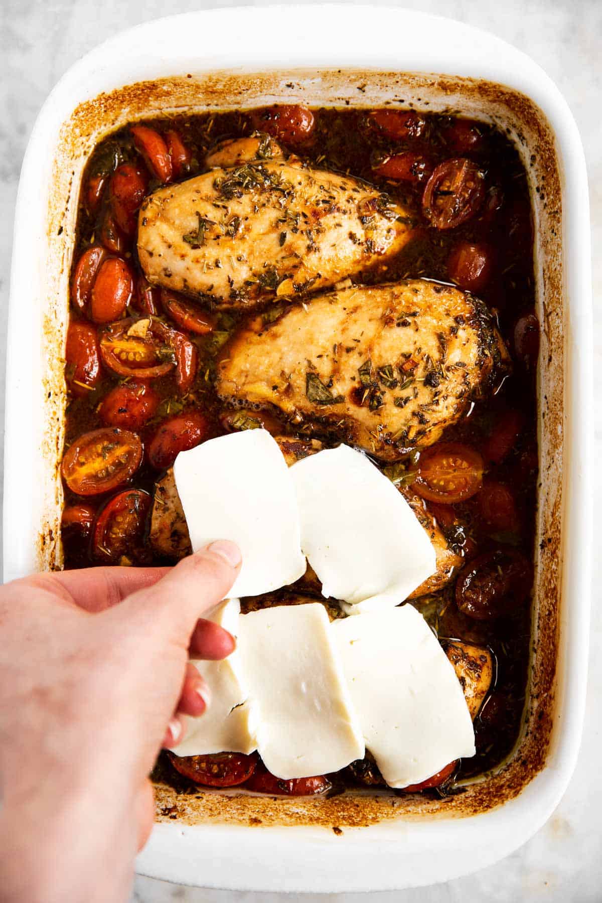 female hand placing slice of mozzarella cheese on baked chicken breast in dish with balsamic and cherry tomatoes