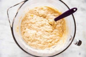 glass mixing bowl with banana pancake batter, with purple rubber spatula stuck in batter