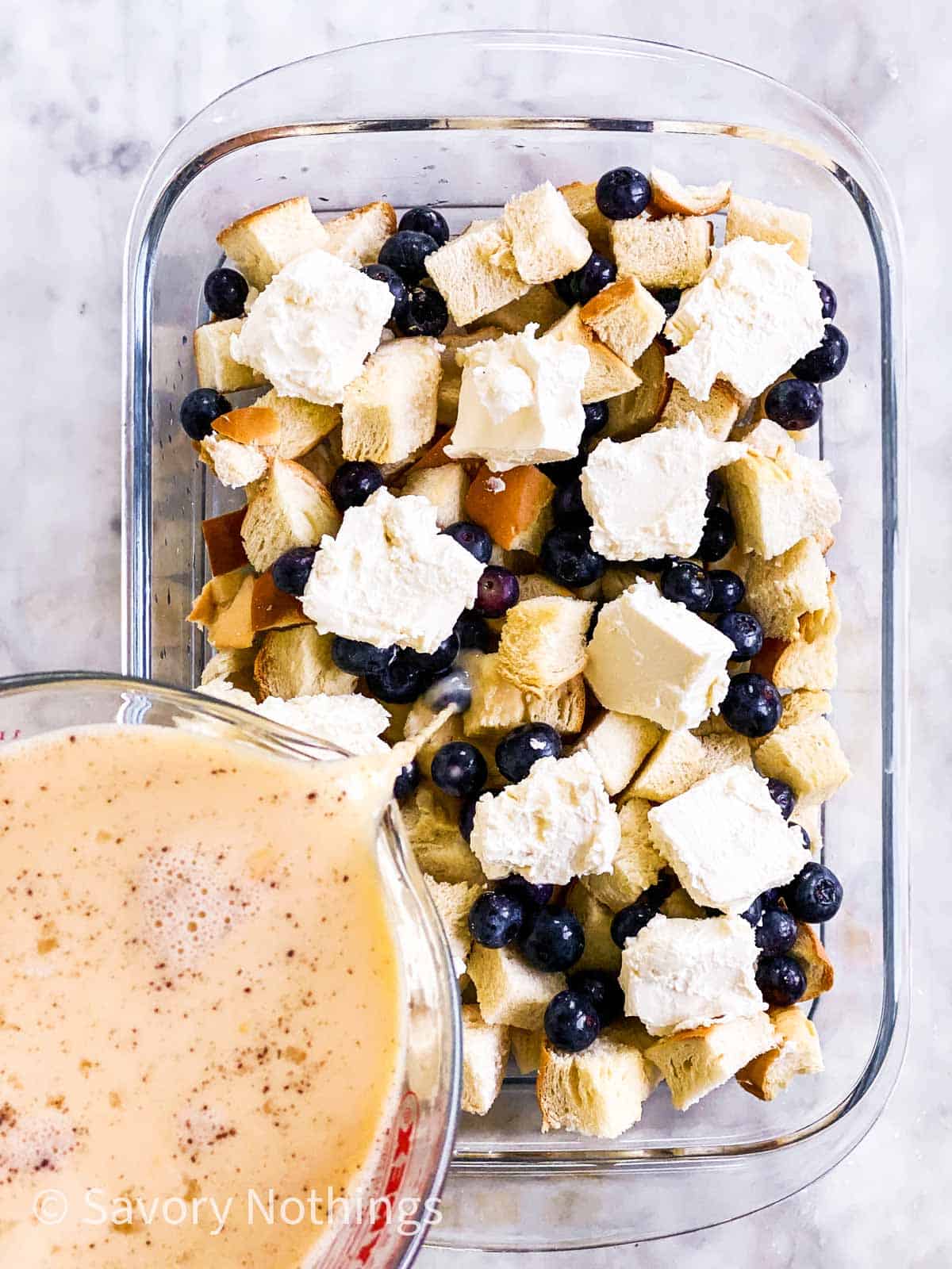 egg and milk mixture pouring over blueberry French toast casserole ingredients in glass dish