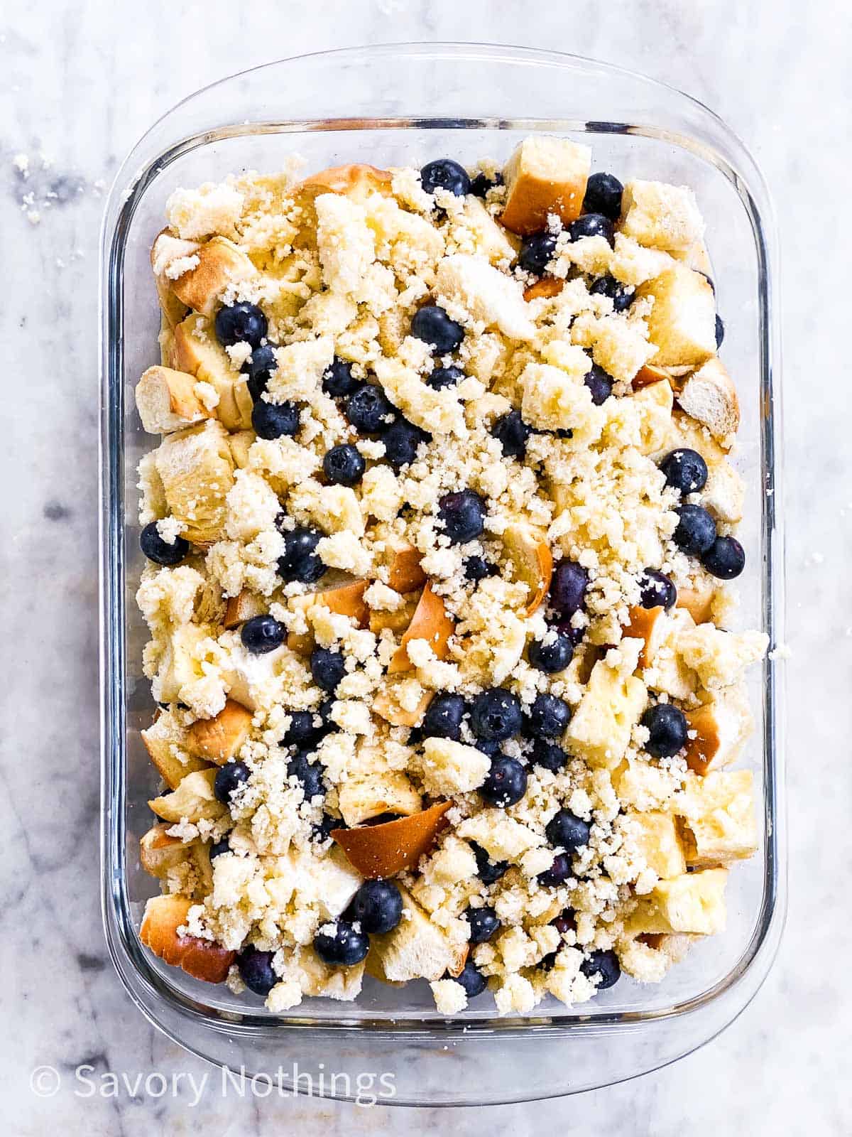 assembled but unbaked blueberry French toast casserole in glass dish sitting on white marble surface, topped with streusel