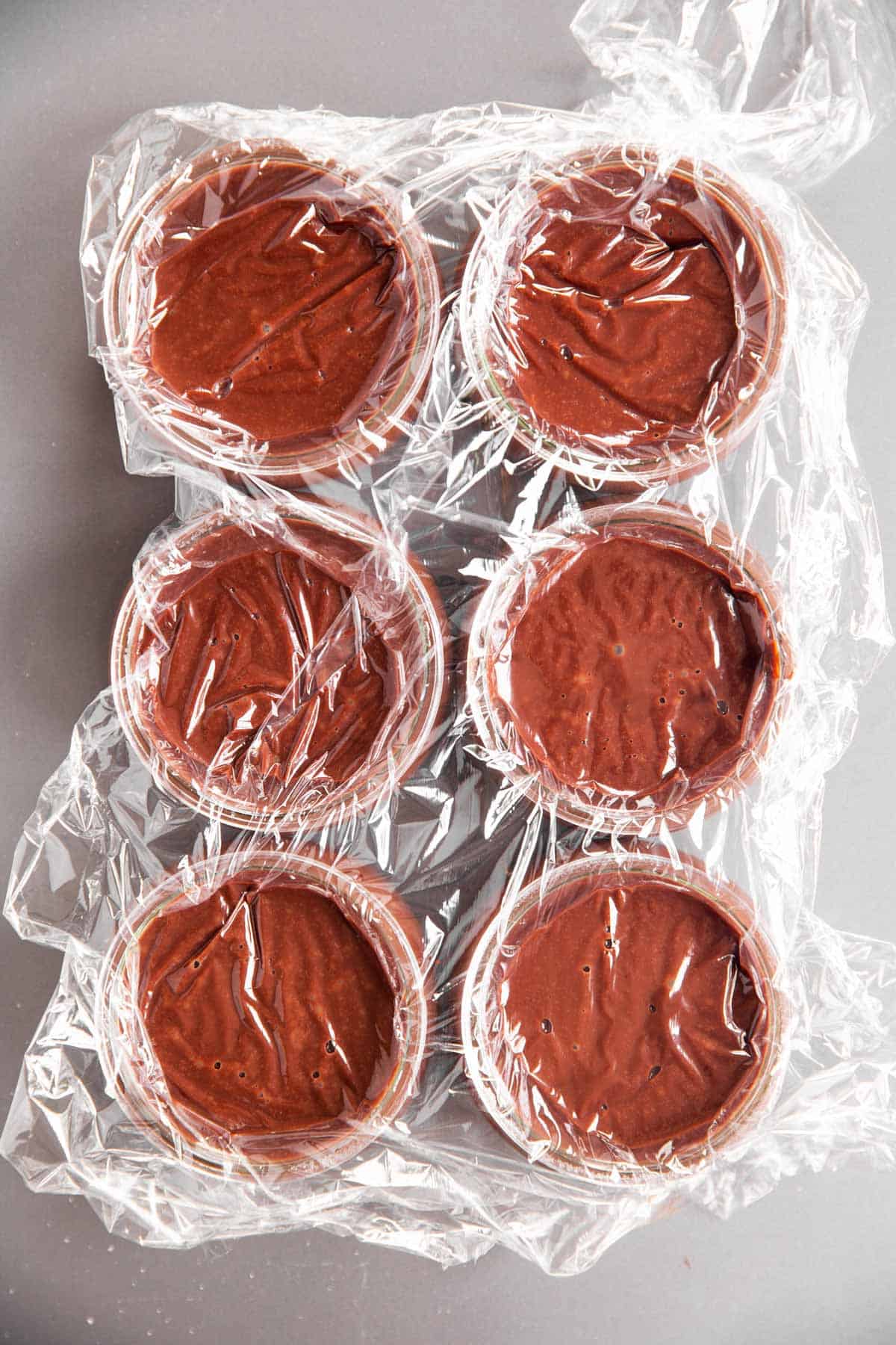six glass jars filled with chocolate pudding, covered with plastic wrap