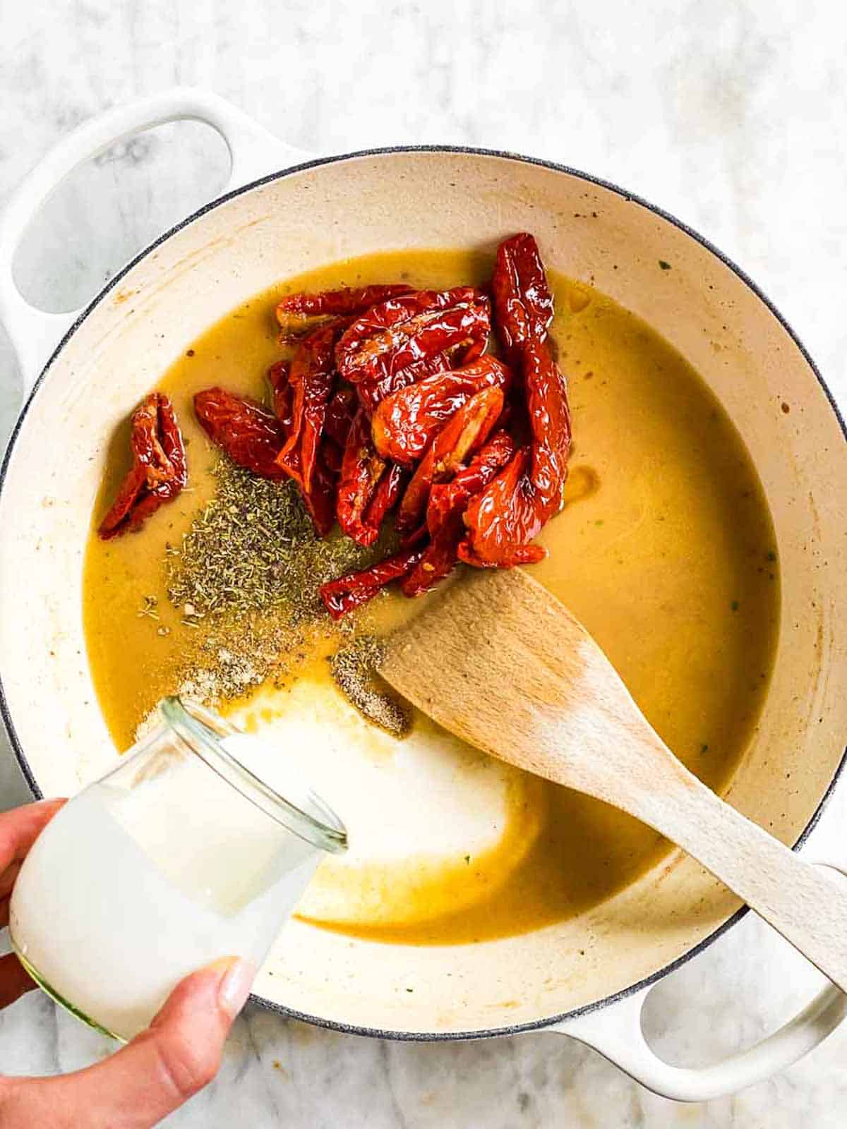 female hand pouring heavy cream into skillet with sauce and sun-dried tomatoes