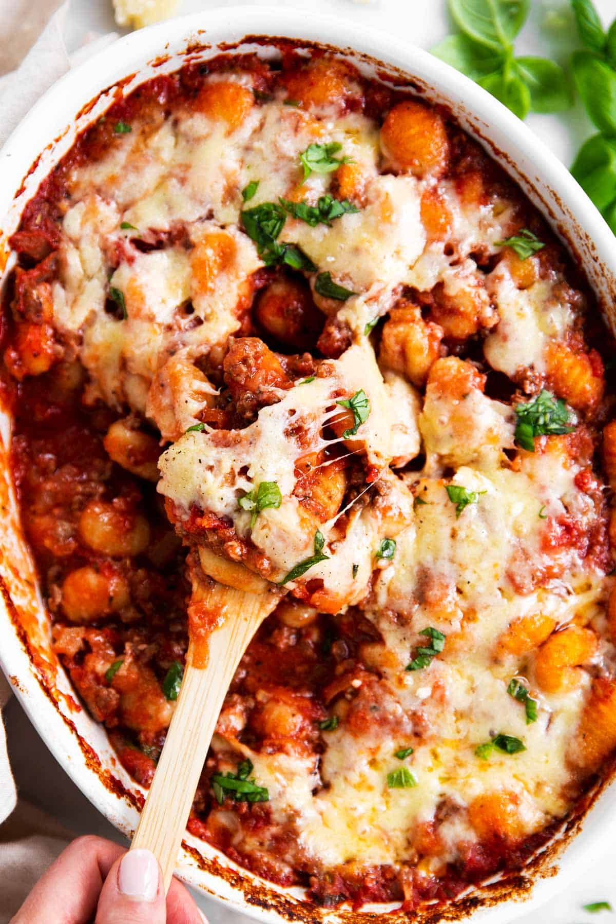 female hand using wooden spoon to scoop gnocchi bake from white casserole dish