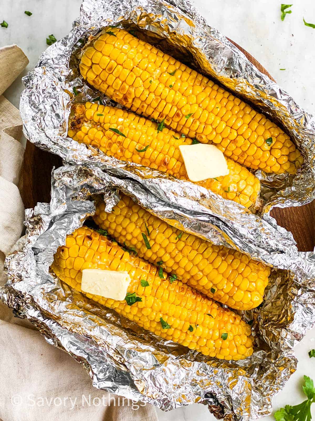 two opened foil packets with two ears of grilled corn each on wooden platter