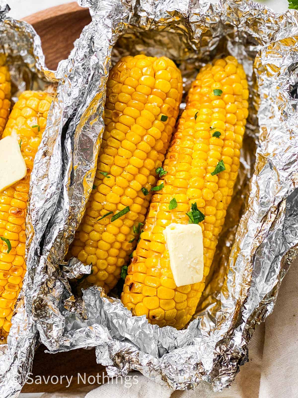 close up photo of two grilled ears of corn in aluminium foil packet with a pat of butter on top