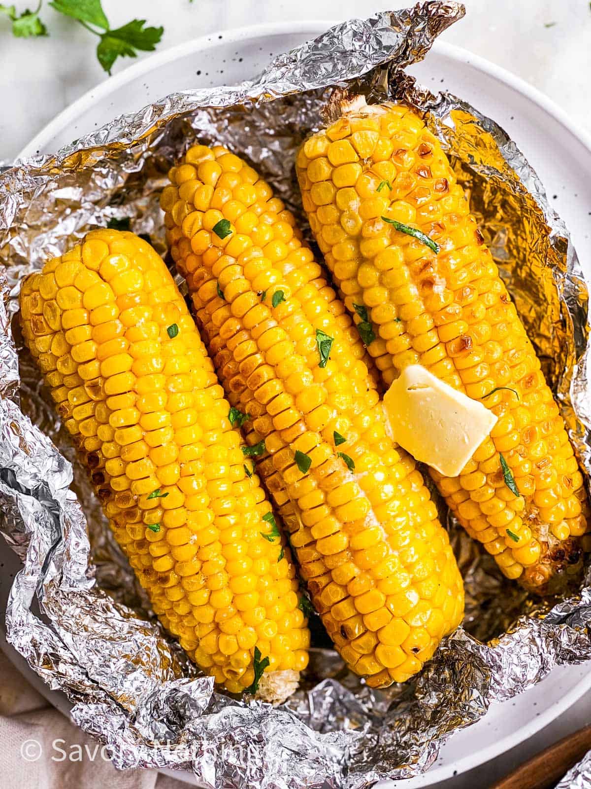 three grilled cobs of corn in opened foil packet on white plate with butter and chopped parsley