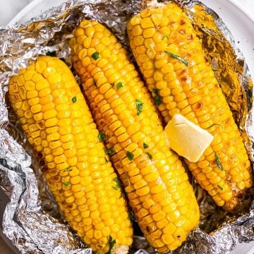 three grilled cobs of corn in opened foil packet on white plate with butter and chopped parsley