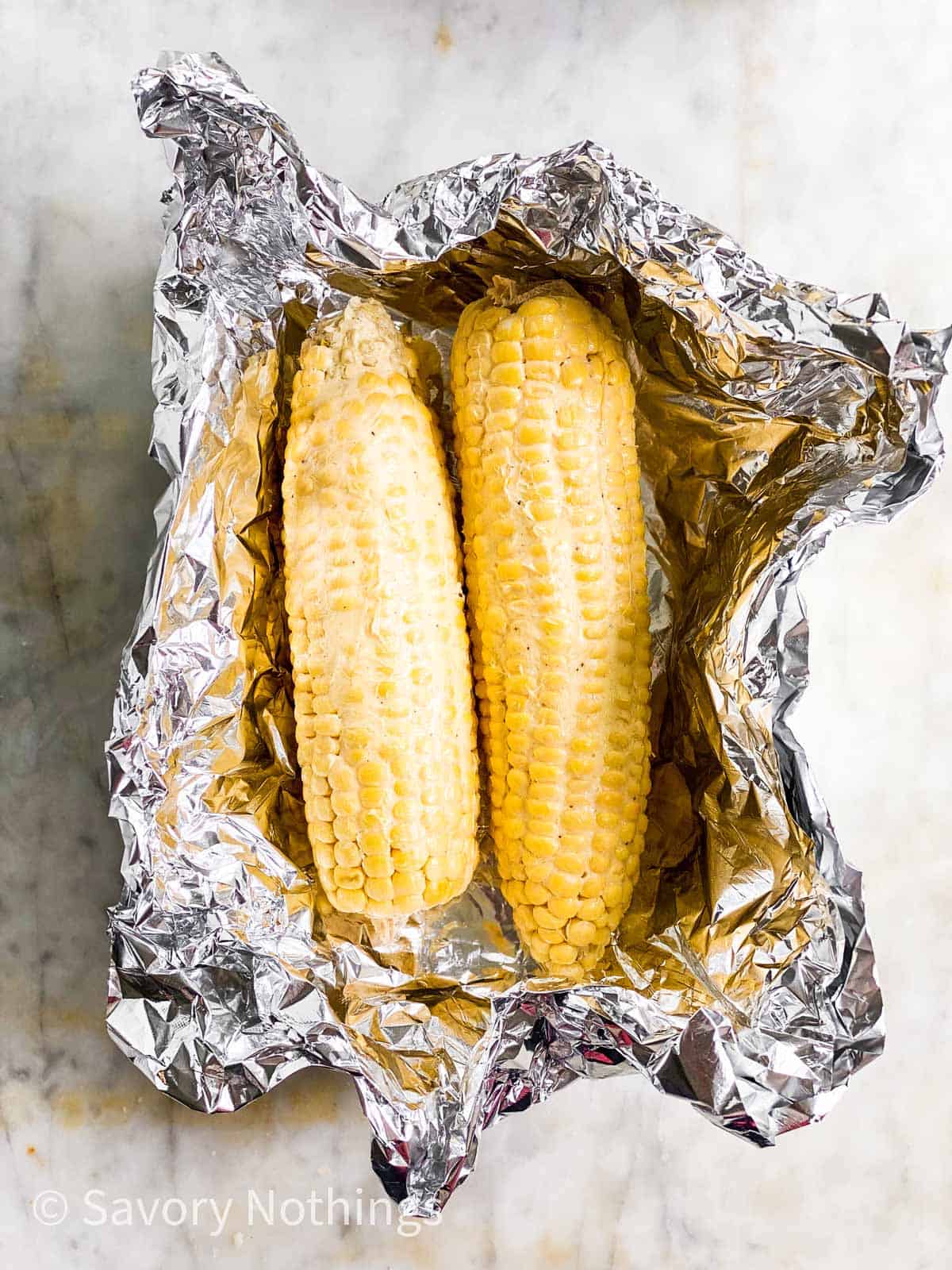 two raw ears of corn covered in butter sitting in aluminium foil boat