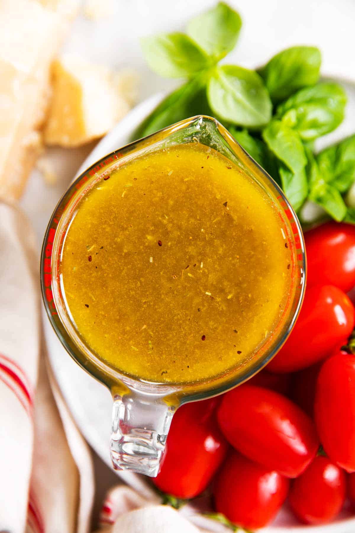 overhead view of italian dressing in glass measuring jug on white plate with tomatoes, basil and parmesan cheese